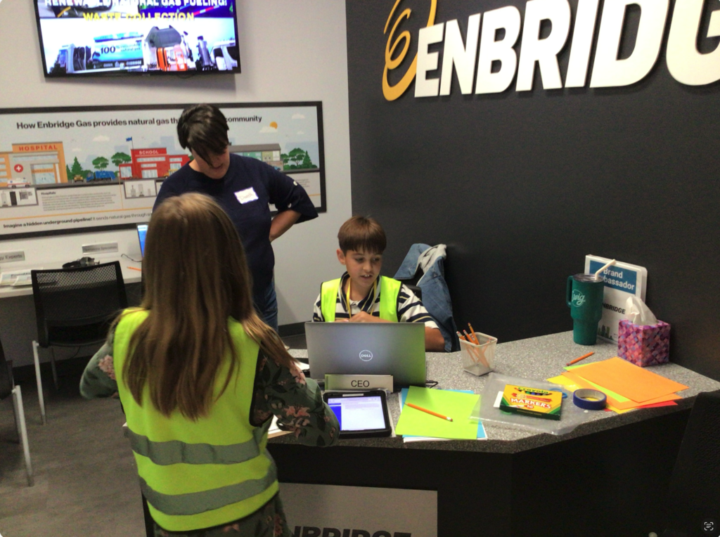 An adult helping a student who is sitting down with a computer in front of him, and a desk sign reading "CEO". Another student is standing in front of the desk back facing the camera with an iPad next to her, some paper, and markers. There is a TV in the back.