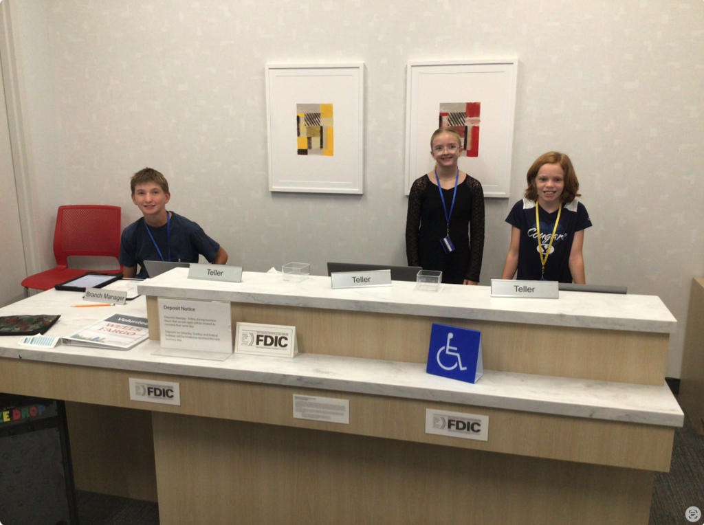 Three students, one sitting on the left with a desk sign saying "Branch Manager". The other two students stand to the right with the desk signs reading "Teller". Two abstract paintings behind them. A blue handicap sign on top of the desk to the right. "FDIC" signs throughout to read the name of the company.