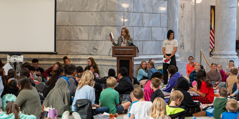 A group of young people sits on the floor in the State Capitol rotunda, attentively listening as Utah's First Lady Abby Cox speaks from a podium