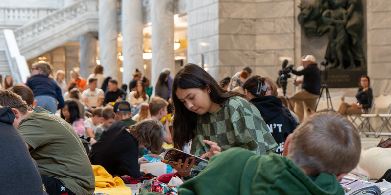 A group of people, including children and adults, are seated on blankets in the Utah State Capitol , reading books and enjoying a gathering.