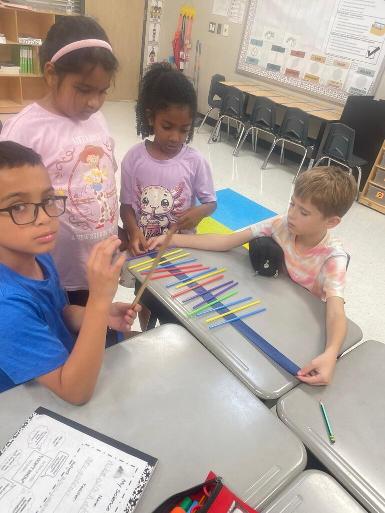 Students working together collaboratively at desks.   they are using tape, rulers, and straws to create sound waves.