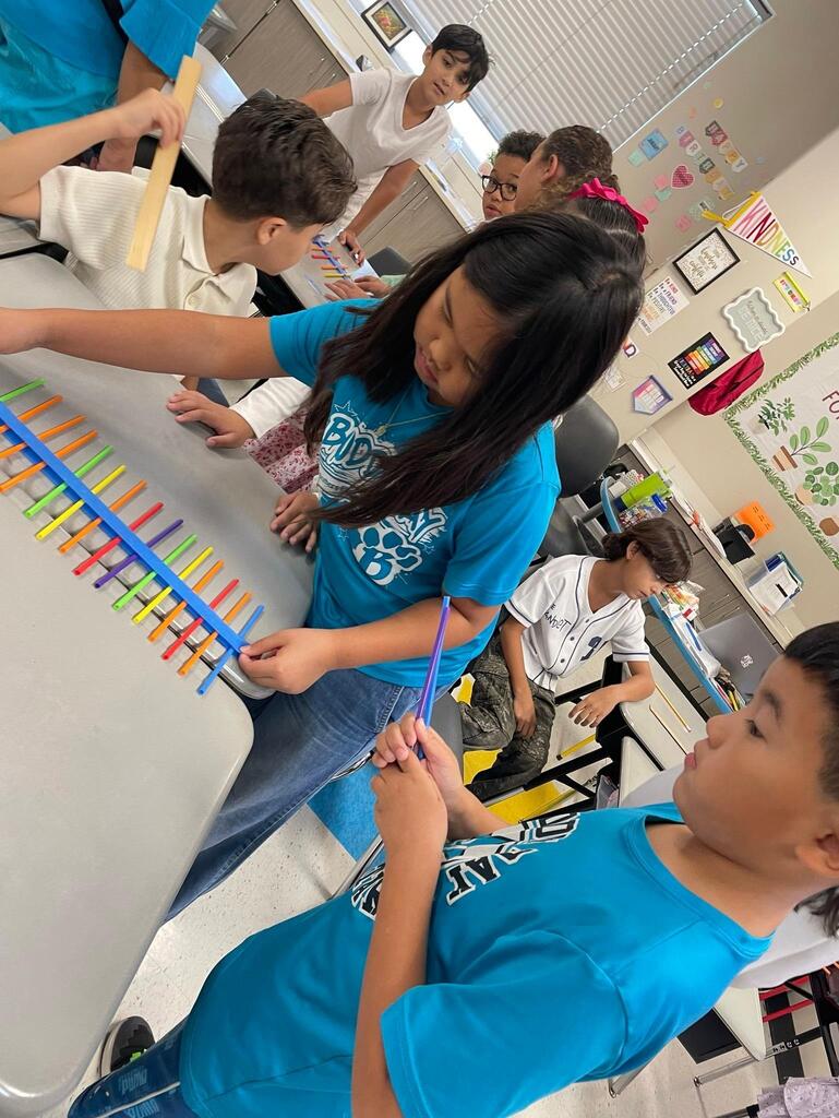 Students working together collaboratively at desks.   they are using tape, rulers, and straws to create sound waves.