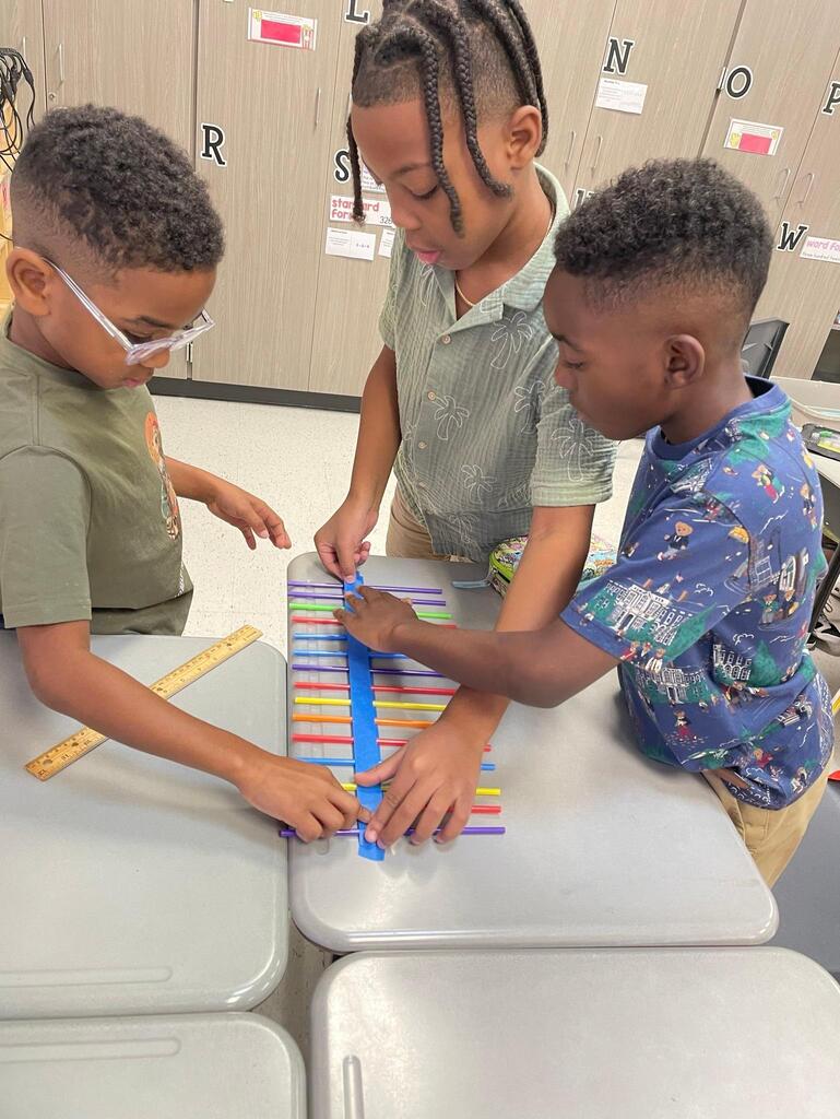 Students working together collaboratively at desks.   they are using tape, rulers, and straws to create sound waves.