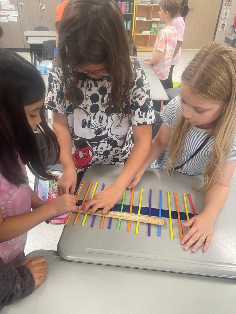 Students working together collaboratively at desks.   they are using tape, rulers, and straws to create sound waves.