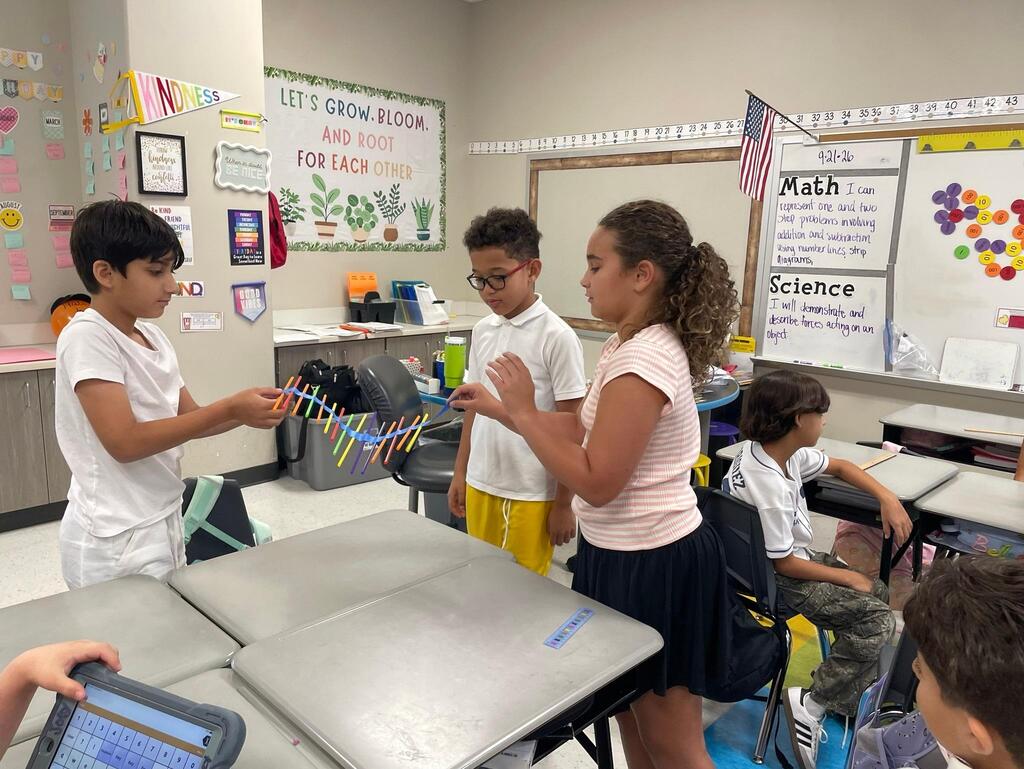 Students working together collaboratively at desks.   they are using tape, rulers, and straws to create sound waves.