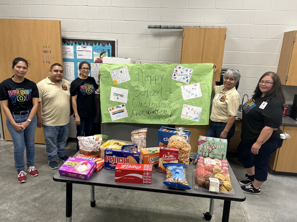 Five school custodial staff members pose together inside a school facility while holding a large, handmade lime green banner. The banner features handwritten text reading "Happy School Custodian Appreciation Day!" along with colorful cards and drawn artwork.