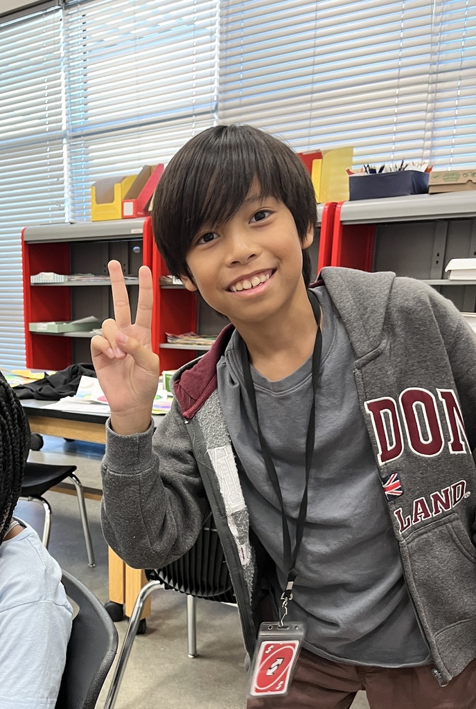 A middle school student smiling and giving a peace sign at his classroom desk while holding an art project.