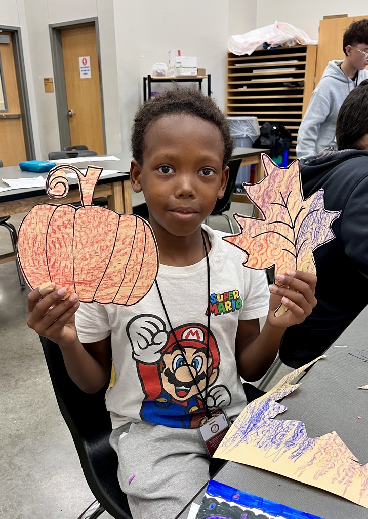 A young middle school student in a graphic shirt holding up his textured pumpkin and leaf artwork in a classroom.