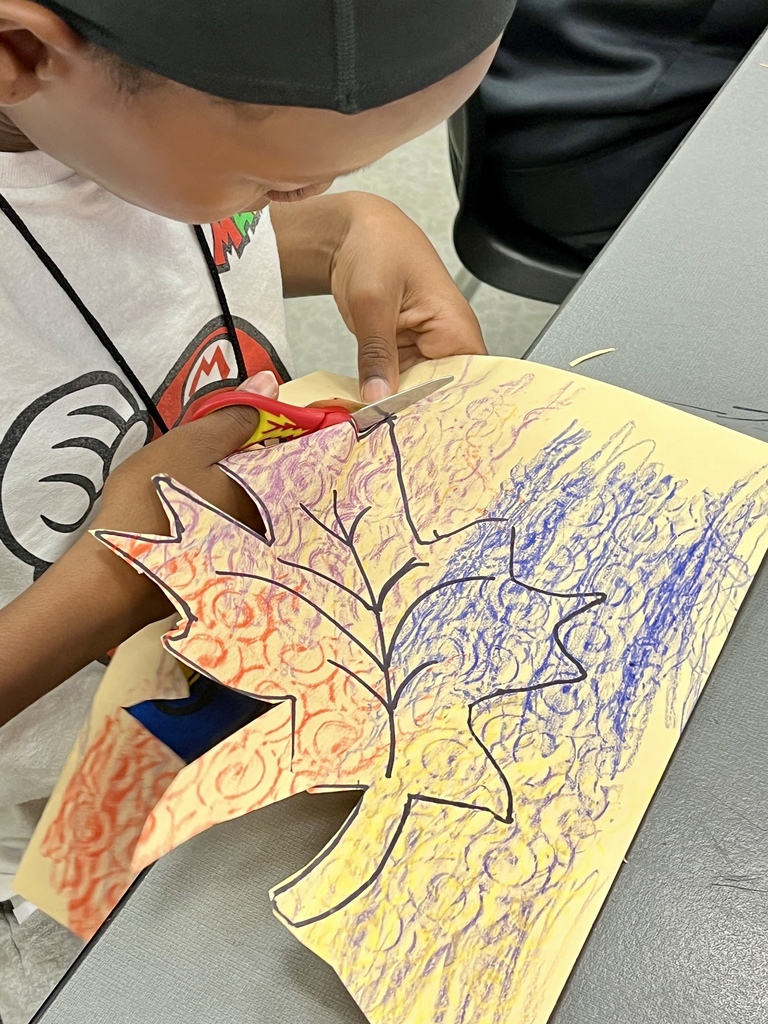 A student using scissors to carefully cut along the outline of a colorful, textured crayon leaf drawing.