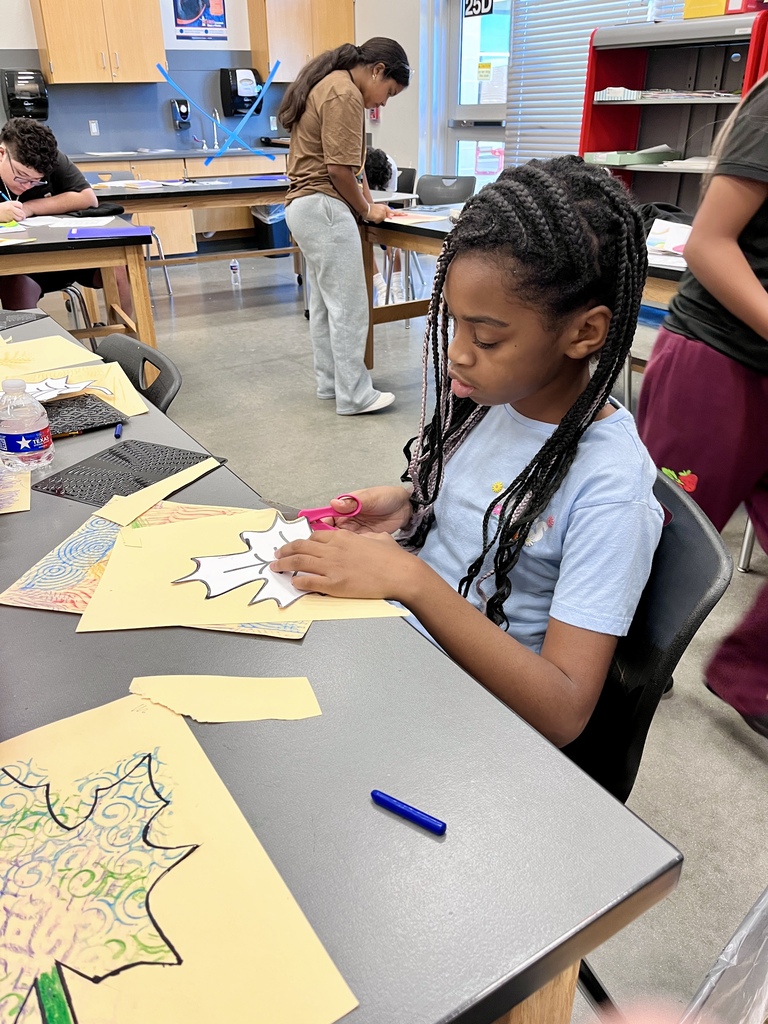A middle school student focused on carefully cutting out leaf patterns at her desk in an art classroom.