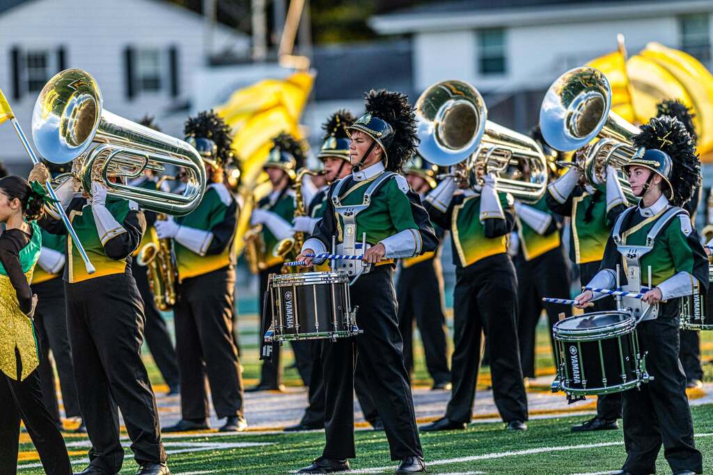 Amherst Marching Comets percussionists and brass musicians perform on the field in green, gold, black and white uniforms while yellow color guard flags wave in the background.