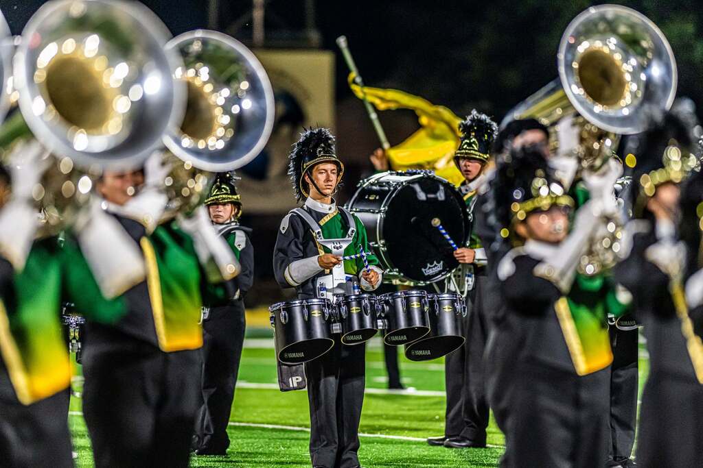 Members of the Amherst Marching Comets perform under the stadium lights in green, gold, black and white uniforms. A student plays tenor drums at the center while brass musicians and a yellow color guard flag frame the scene.