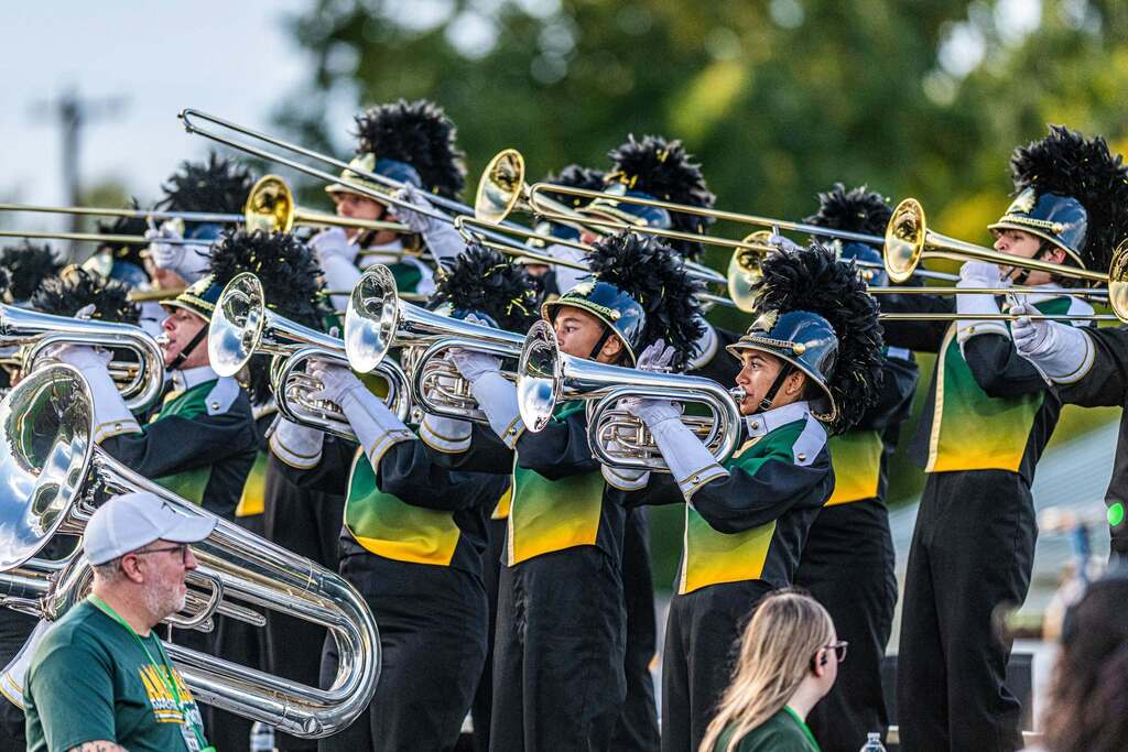 Members of the Amherst Marching Comets brass section perform outdoors in green, gold, black and white uniforms, playing baritones, trombones and tubas.