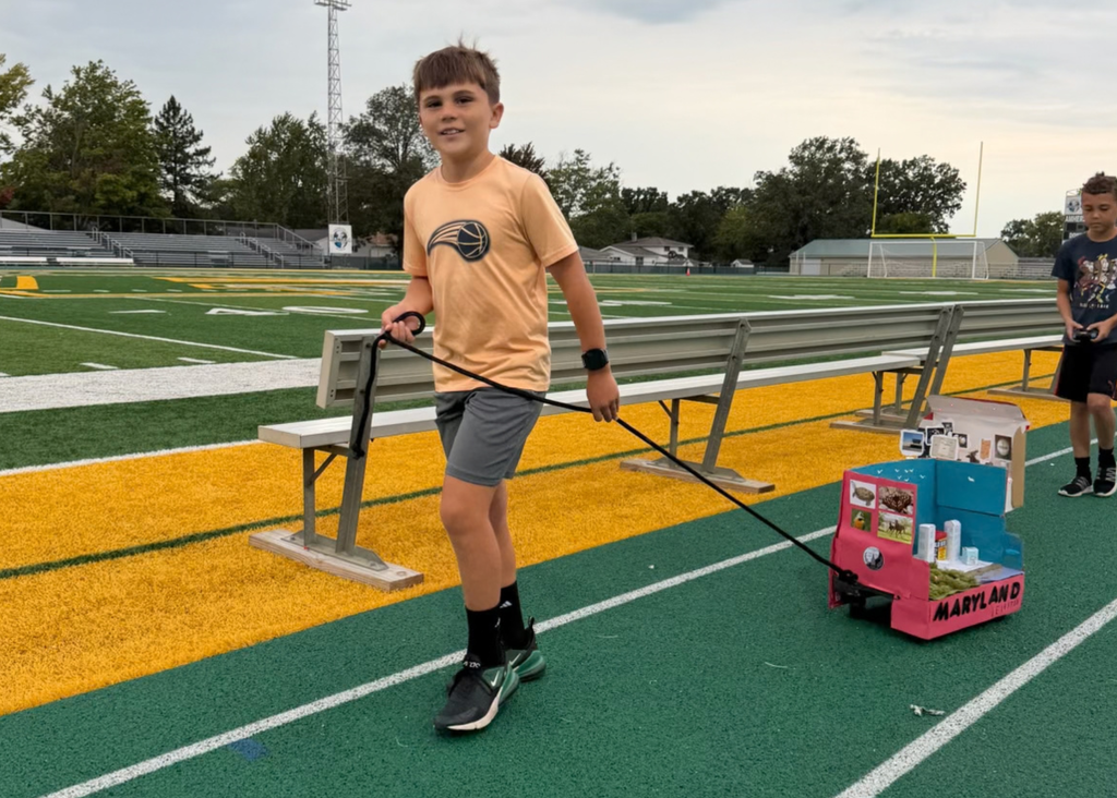 A student in an orange shirt pulls a pink-and-blue Maryland float decorated with wildlife pictures and miniature buildings along the track.