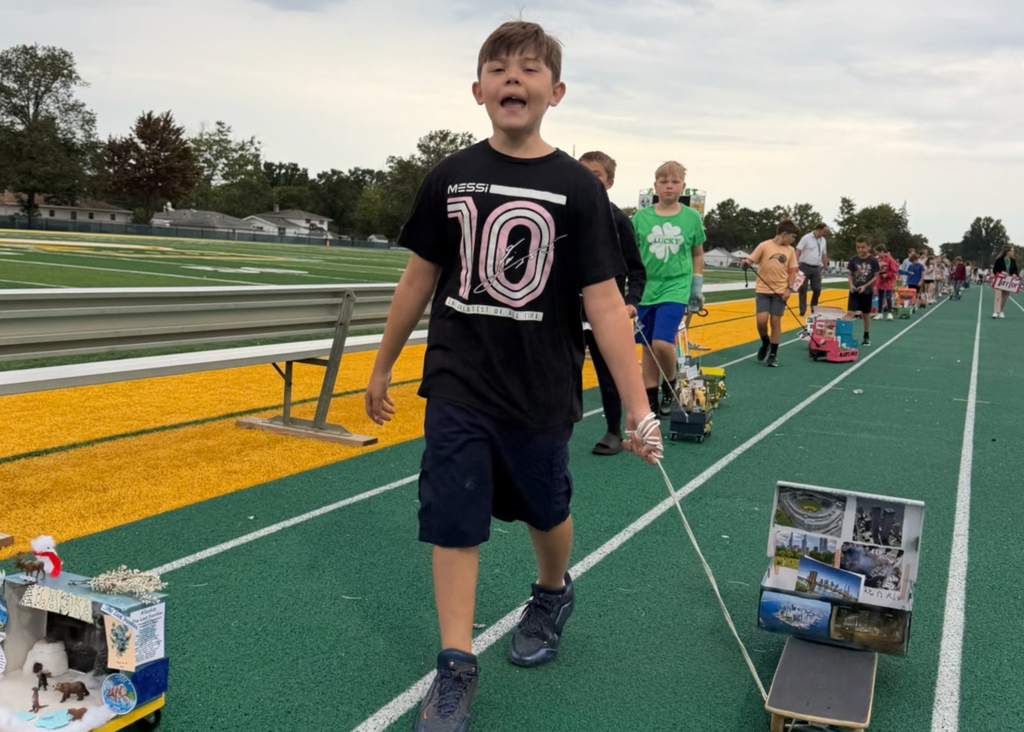 A student in a black Messi shirt pulls a handmade float along the track, followed by a line of classmates with their state projects.