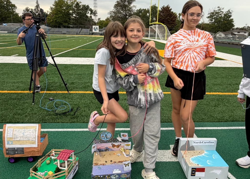 Three students pose on the athletic field beside handmade state floats, including a North Carolina display with a model coastline. A student operates a video camera in the background.