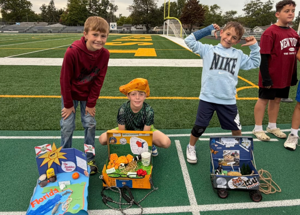 Three students pose behind Florida, Wisconsin, and Idaho floats decorated with state symbols. The student beside Wisconsin wears a cheese-shaped hat.