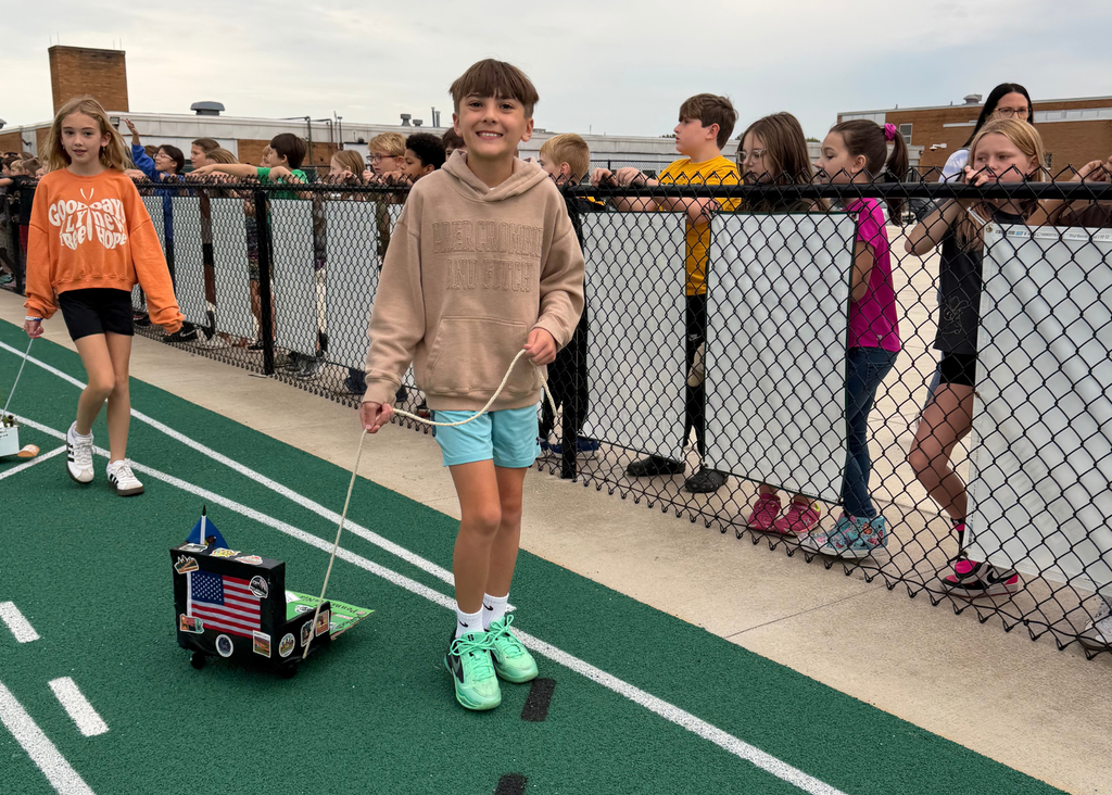 A smiling student in a tan hoodie pulls a black float decorated with an American flag and state images while classmates watch from behind the fence.