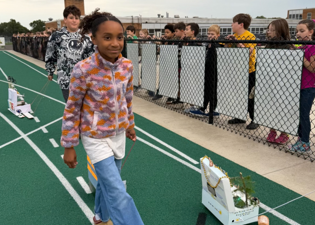 A student in a colorful fleece jacket pulls a white state float decorated with beads and miniature trees while classmates watch from behind the fence.