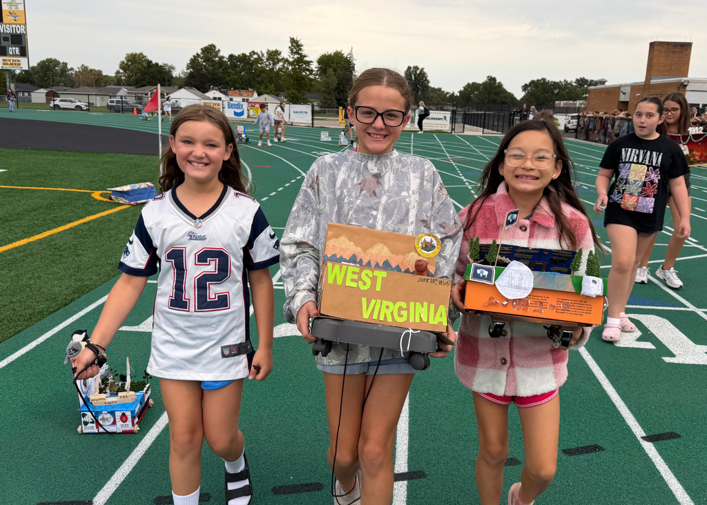 Three smiling students display state projects on the track, including a Boston Tea Party scene, a West Virginia mountain display, and a Wyoming float.