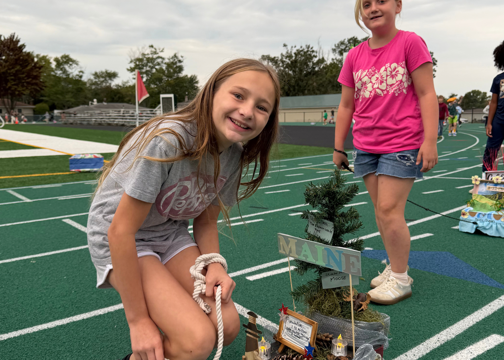 Two students pose beside a Maine float featuring a miniature pine tree, pinecones, a moose figure, and state facts.