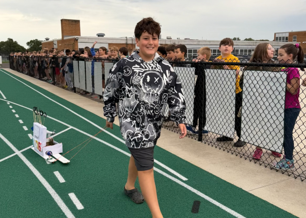 A smiling student in a black-and-white hoodie pulls a handmade state float along the track as classmates watch from behind the fence.