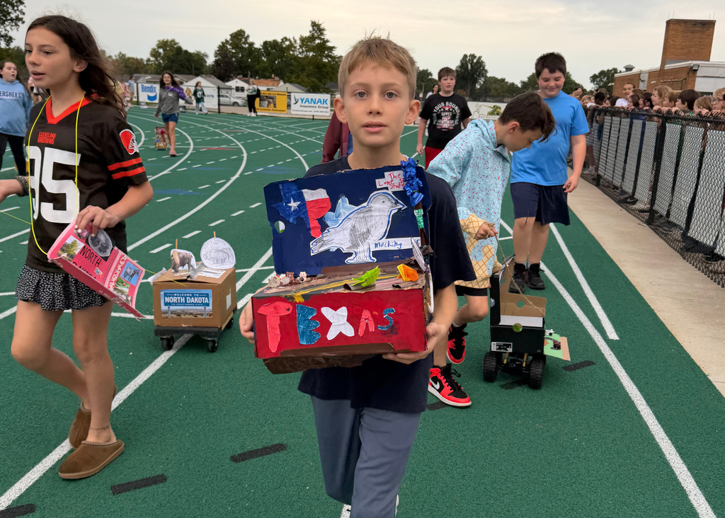 A student holds a colorful Texas project featuring the state outline, flag, and a drawing of a mockingbird. Classmates walk nearby with other state floats.