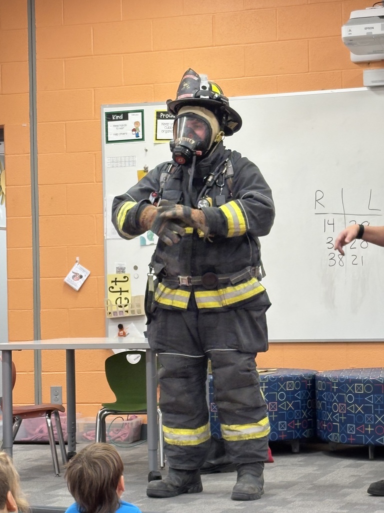 An Amherst firefighter demonstrates full protective gear, including a helmet, face mask, gloves, and reflective clothing, as students watch in a Powers Elementary classroom.