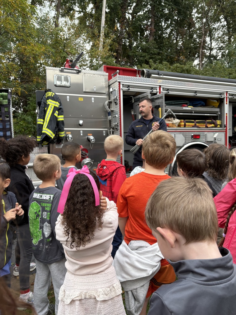 An Amherst firefighter holds a long metal tool while speaking to Powers Elementary students gathered beside a fire truck. Open compartments reveal firefighting equipment.