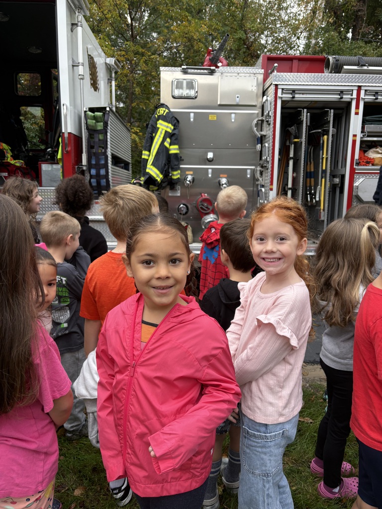 Powers Elementary students gather beside a fire truck with open equipment compartments. Two students smile at the camera, with a firefighter’s protective coat and tools displayed behind them.