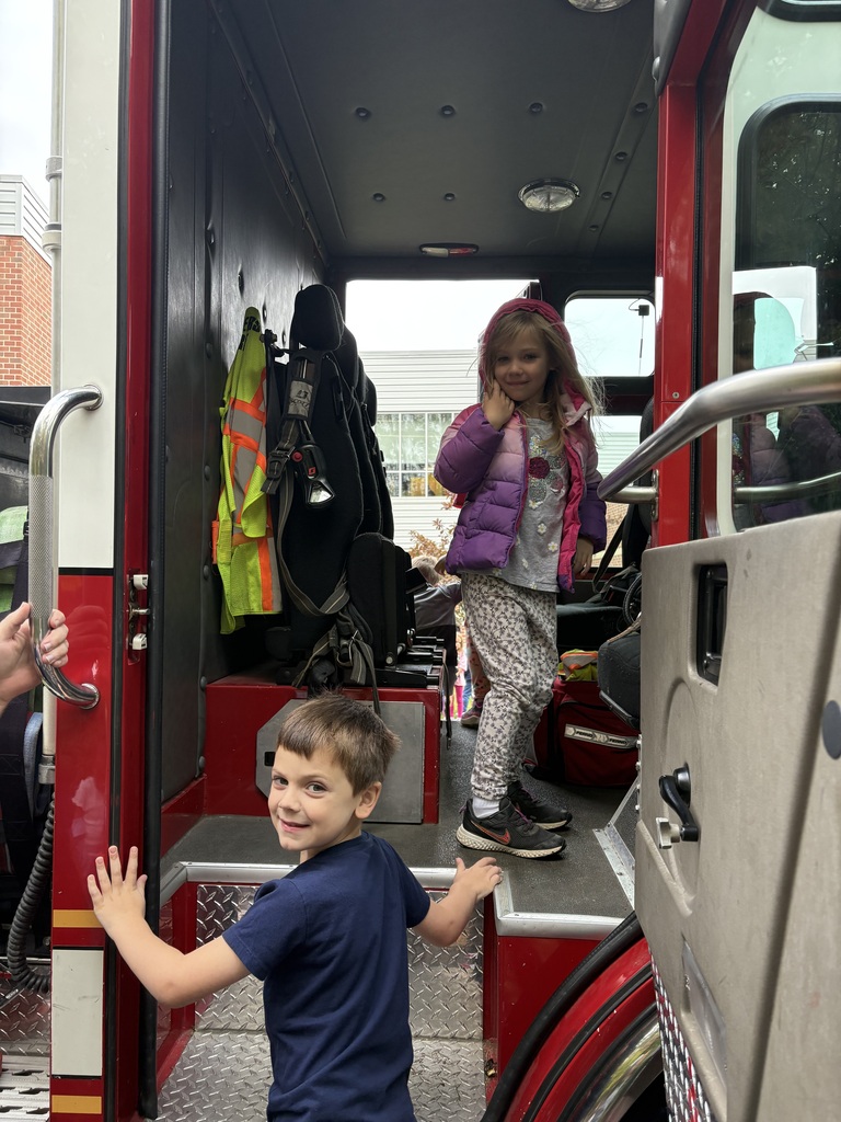 Two Powers Elementary students explore the cab of a fire truck. One stands inside beside firefighter equipment while the other smiles at the camera from the entrance steps.