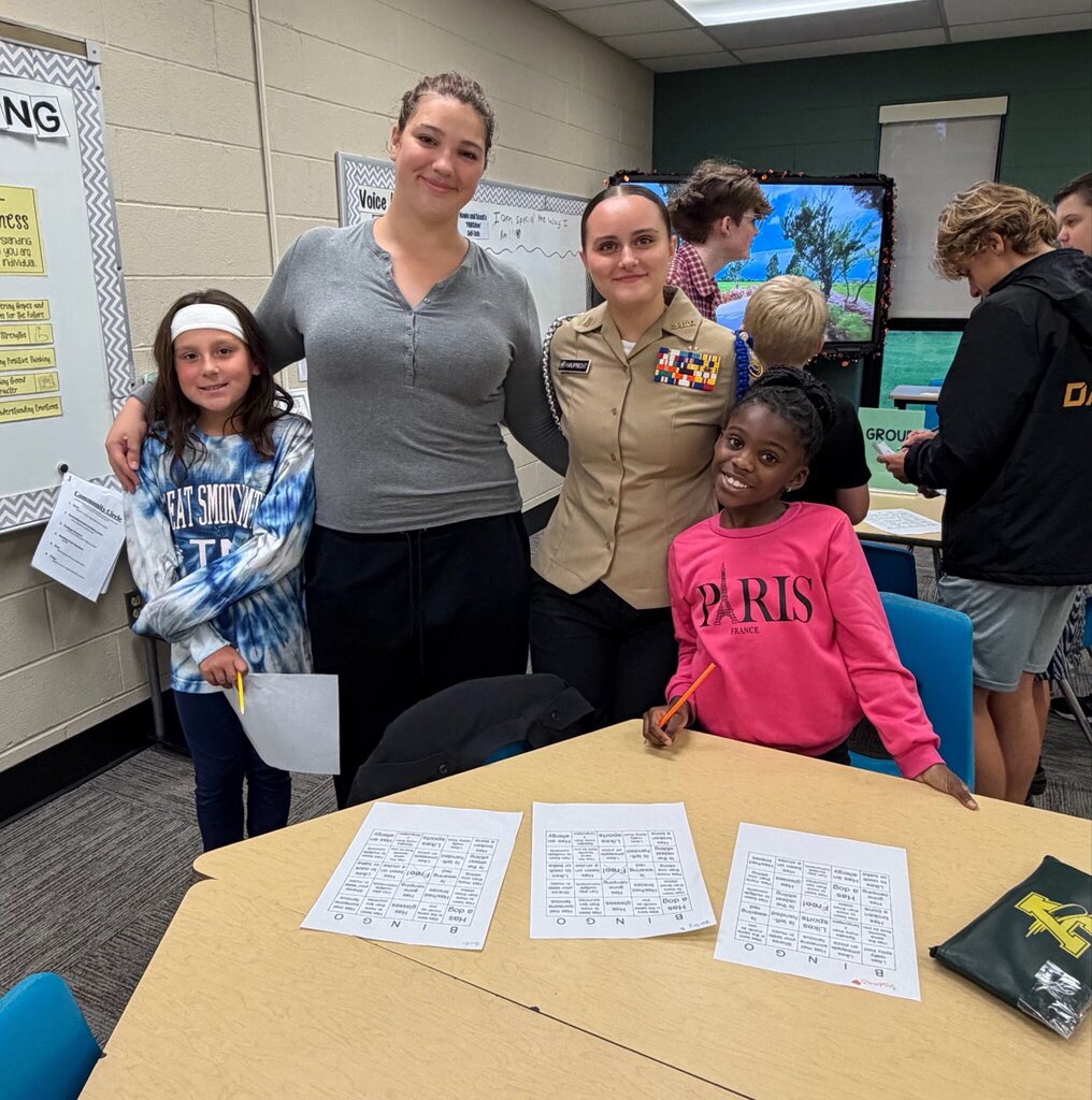 Two younger Comets pose with two older mentors, including a student in a Navy cadet uniform, during the first Marion Mentoring session. The group gathers around a table with bingo sheets as other students participate nearby.