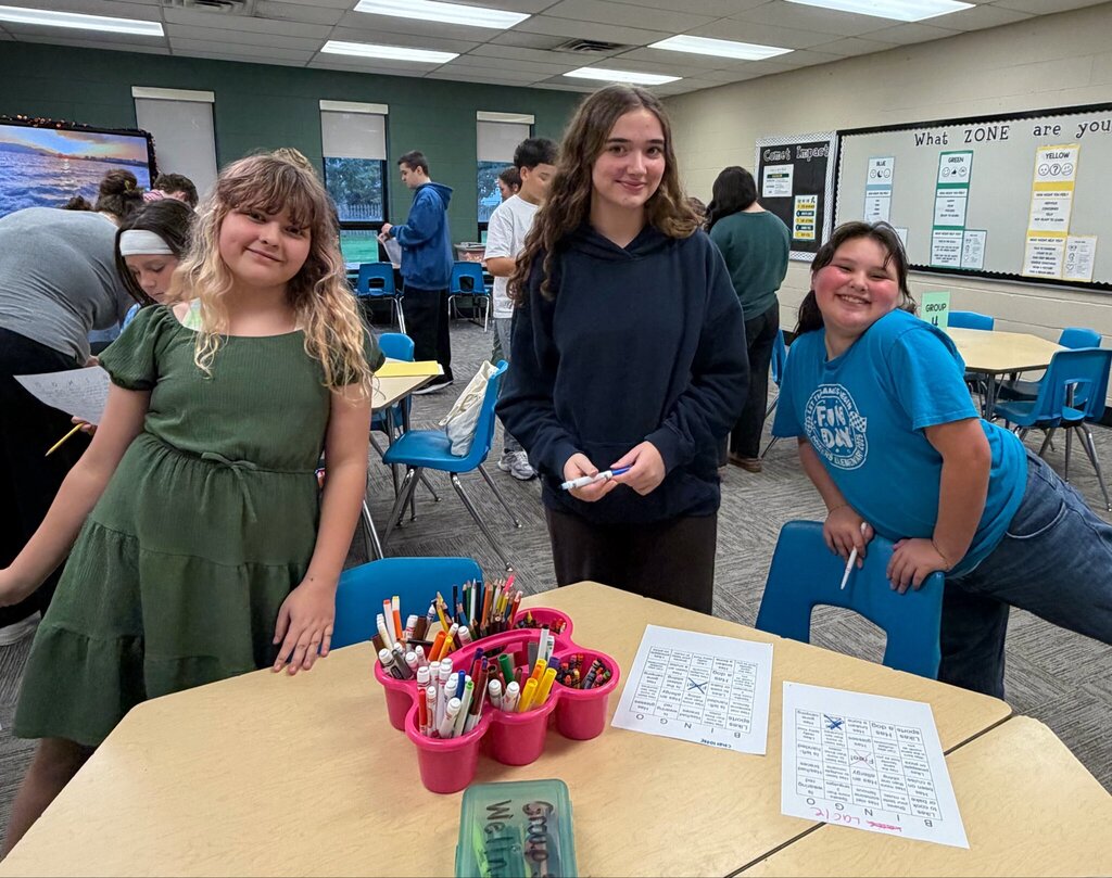 Three smiling students pose together during the first day of Marion Mentoring at Nord Elementary. Activity sheets, markers, and crayons are spread across the table as other students participate in the background.