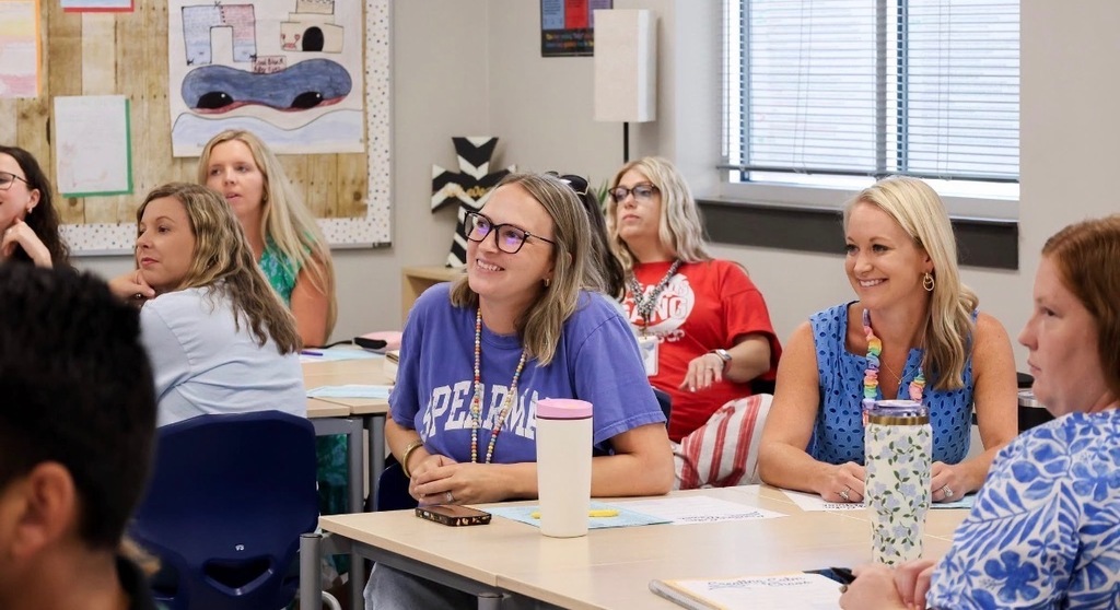 Elementary school teachers participate in a professional development session, seated at tables and listening attentively during a back-to-school training event in a classroom.