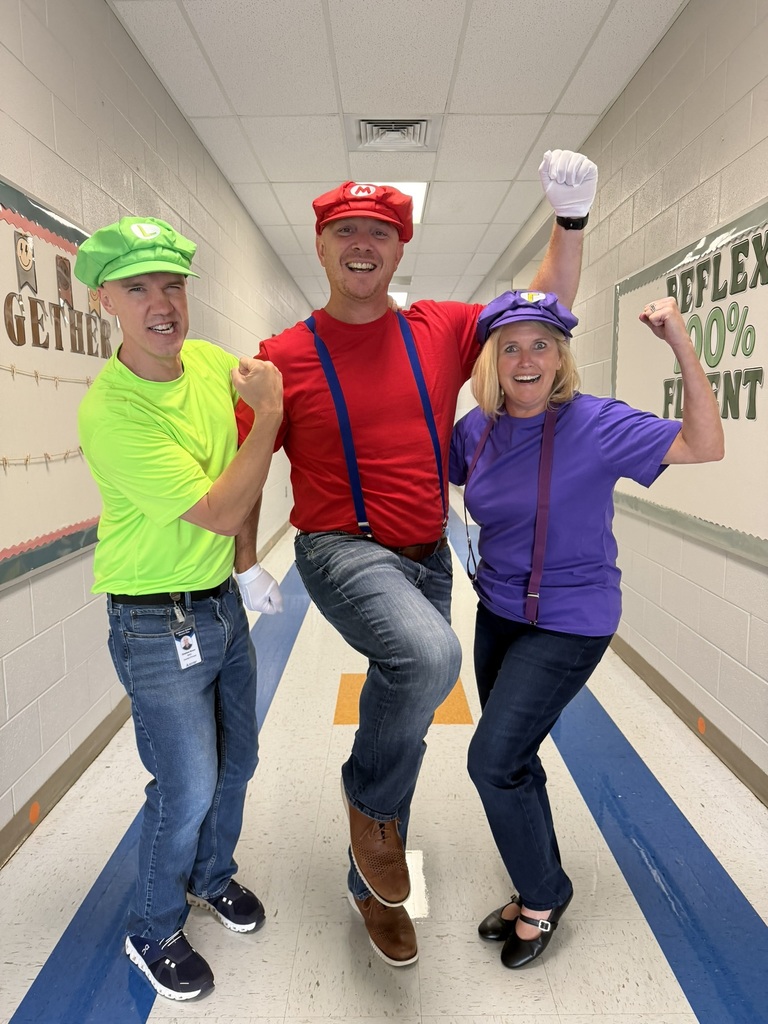 Our 3 admin are posing in colorful video game-inspired costumes pose enthusiastically in a school hallway during a themed school event.