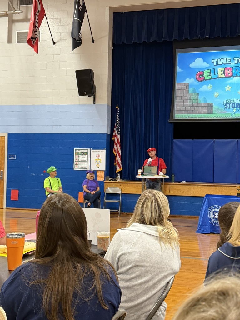 A presenter dressed in a red video game-inspired costume speaks into a microphone during an elementary school professional development session. Two other staff members in green and purple costumes sit nearby on stage while educators watch from the audience.