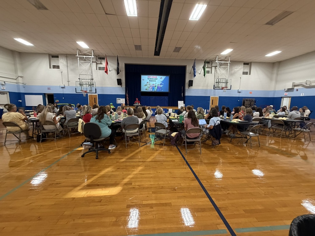 A large group of elementary school staff members sit at tables in a school gymnasium facing a stage, where a presenter leads a professional development session. A presentation is displayed on a large screen as educators prepare for the upcoming school year.