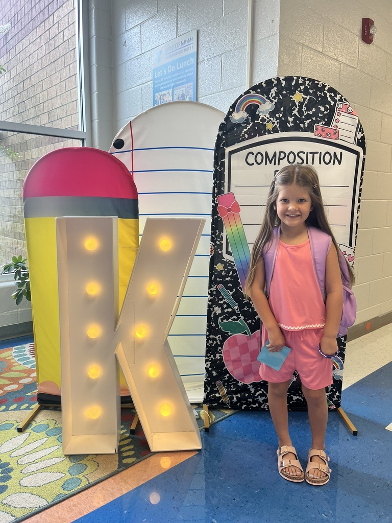A cheerful kindergarten student smiles brightly while holding a "K" sign surrounded by colorful back-to-school decorations like apples, pencils, and a chalkboard background.