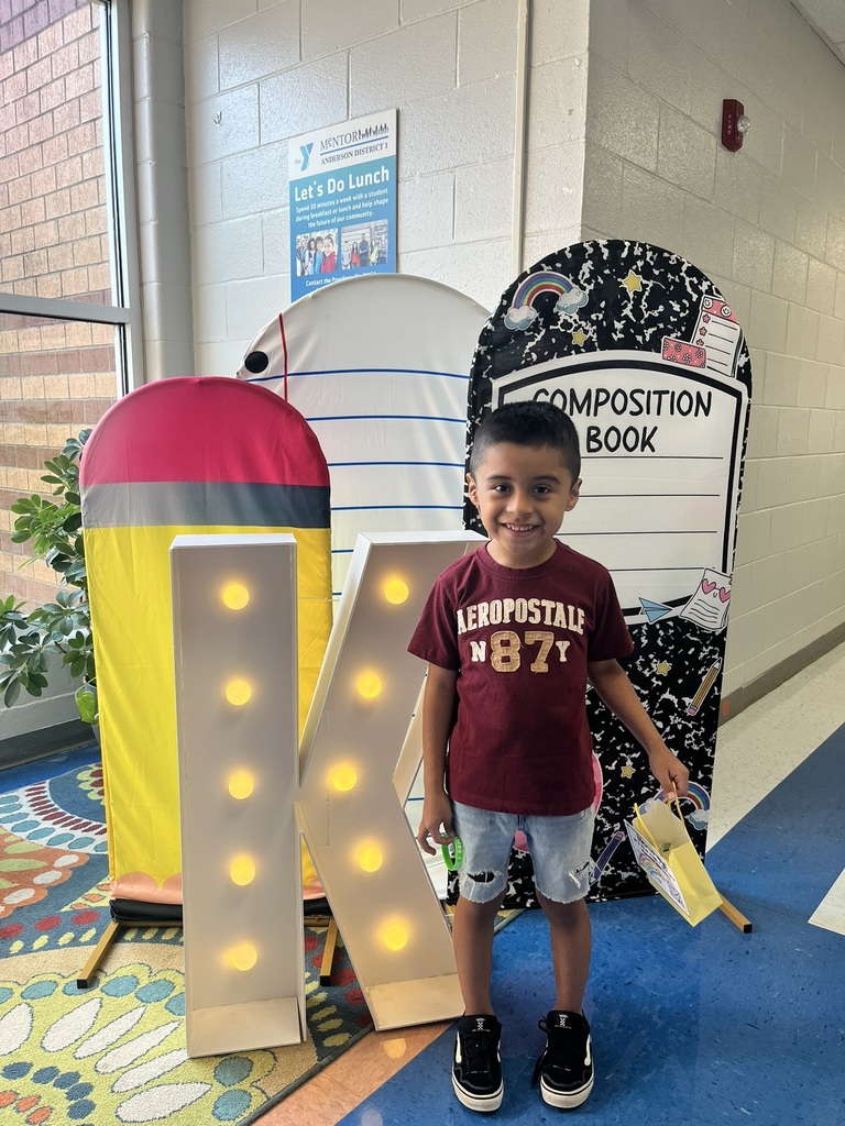 A cheerful kindergarten student smiles brightly while holding a "K" sign surrounded by colorful back-to-school decorations like apples, pencils, and a chalkboard background.