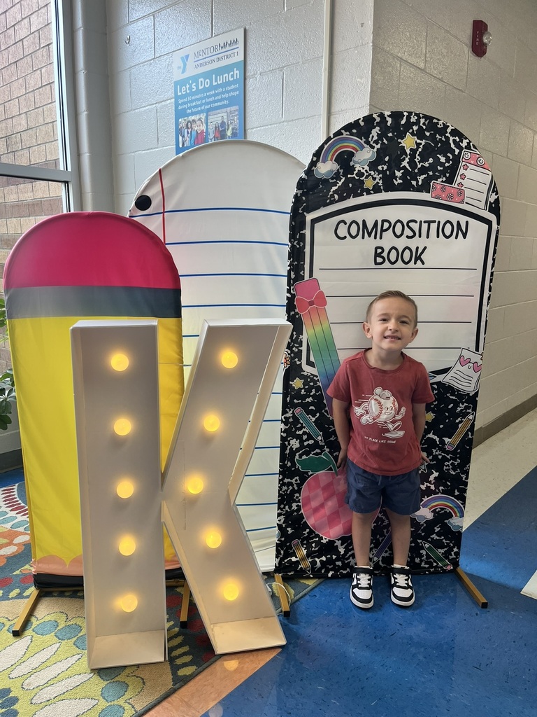A cheerful kindergarten student smiles brightly while holding a "K" sign surrounded by colorful back-to-school decorations like apples, pencils, and a chalkboard background.