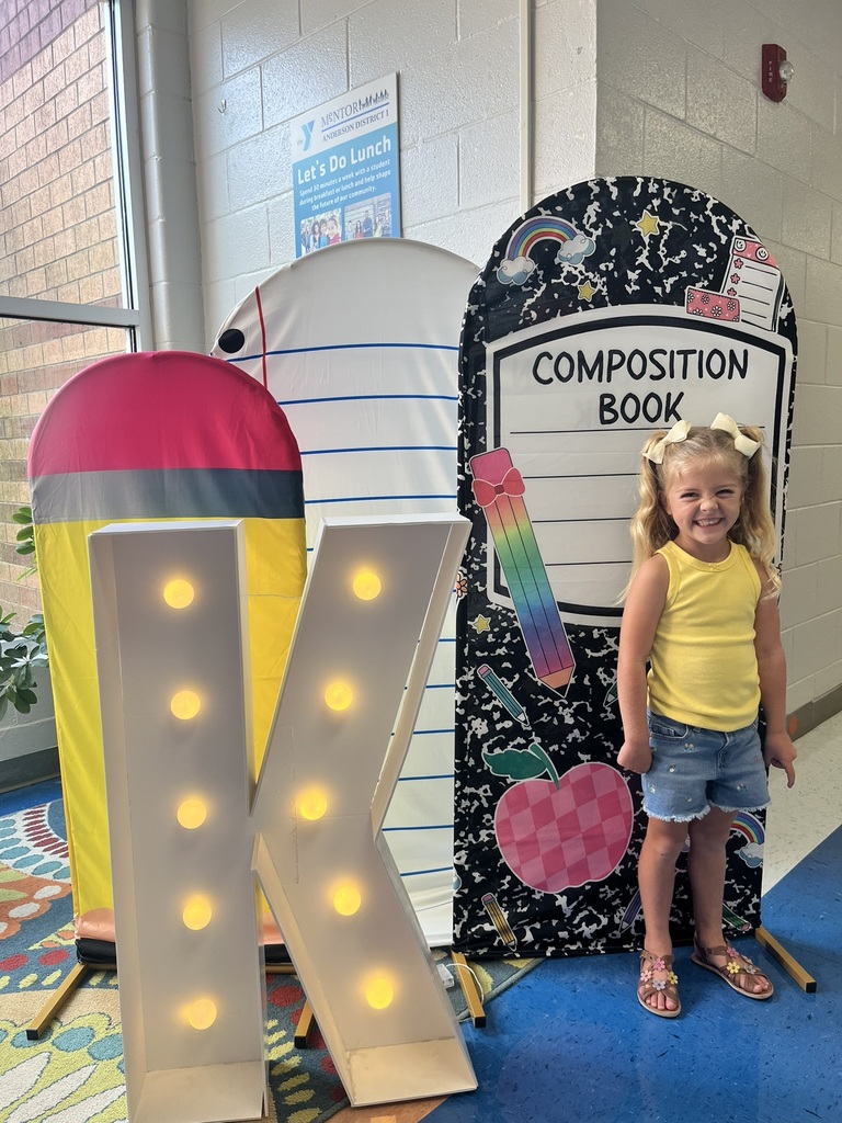 A cheerful kindergarten student smiles brightly while holding a "K" sign surrounded by colorful back-to-school decorations like apples, pencils, and a chalkboard background.