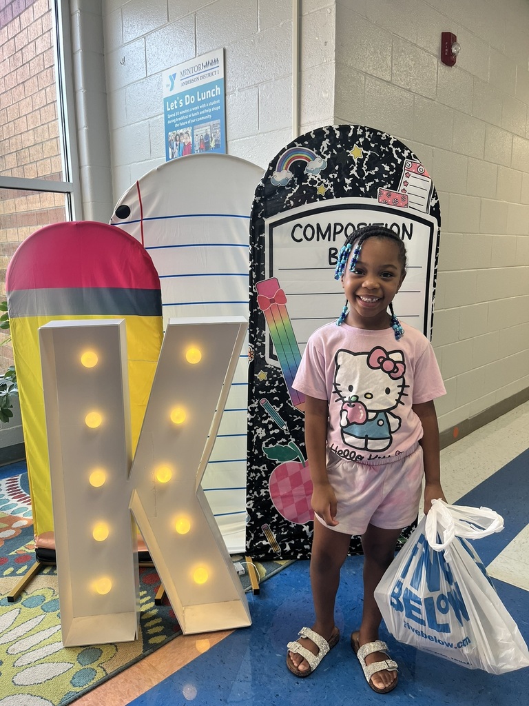A cheerful kindergarten student smiles brightly while holding a "K" sign surrounded by colorful back-to-school decorations like apples, pencils, and a chalkboard background.