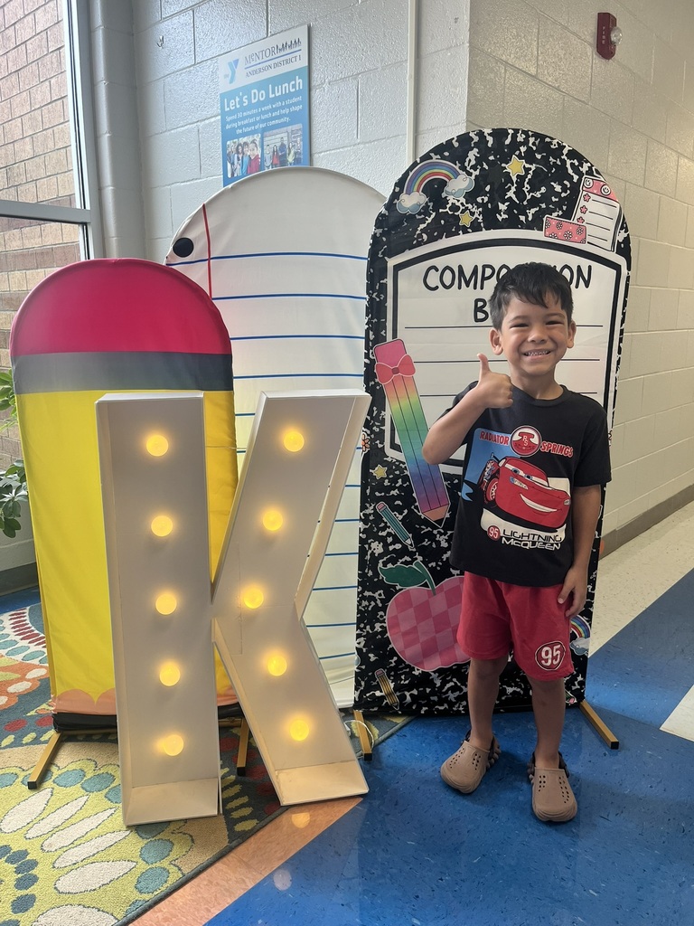 A cheerful kindergarten student smiles brightly while holding a "K" sign surrounded by colorful back-to-school decorations like apples, pencils, and a chalkboard background.
