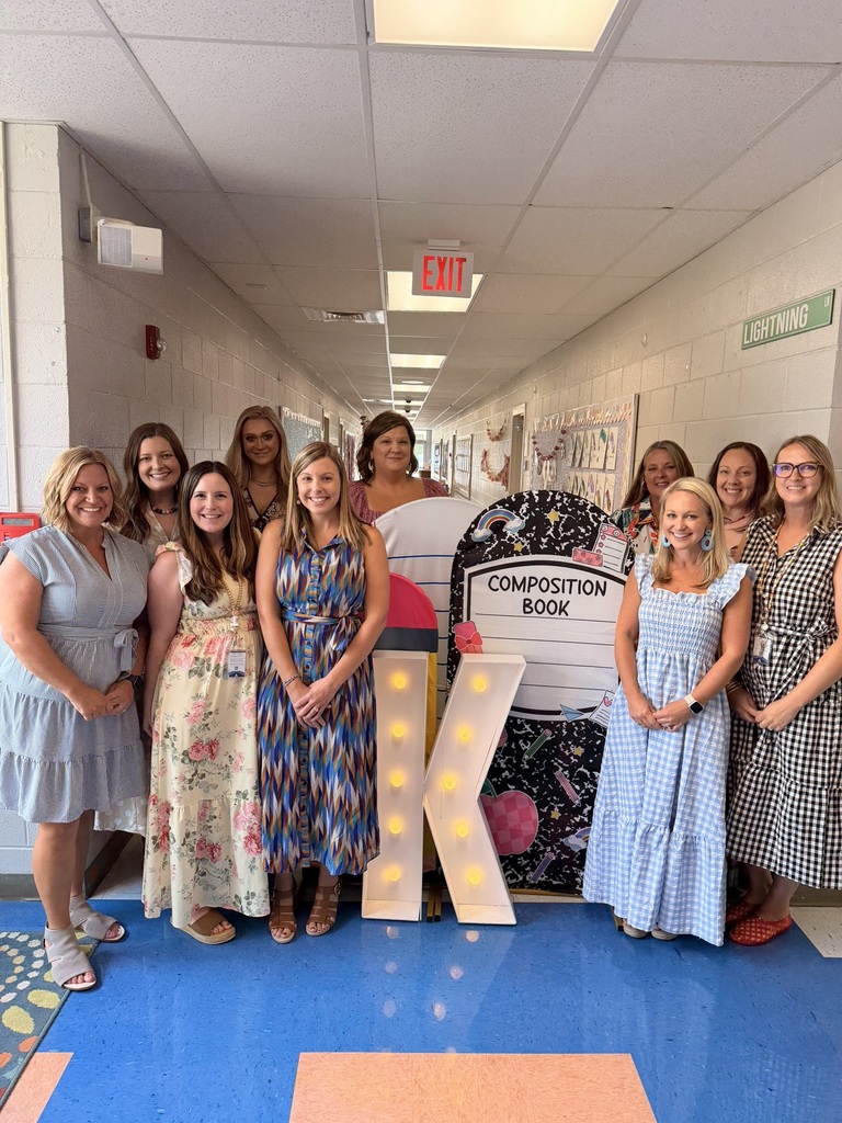 A group photo of kindergarten teachers smiling in front of a school building, each holding up one hand to shape the letter 'K' for kindergarten.