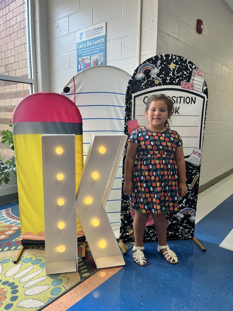 A cheerful kindergarten student smiles brightly while holding a "K" sign surrounded by colorful back-to-school decorations like apples, pencils, and a chalkboard background.