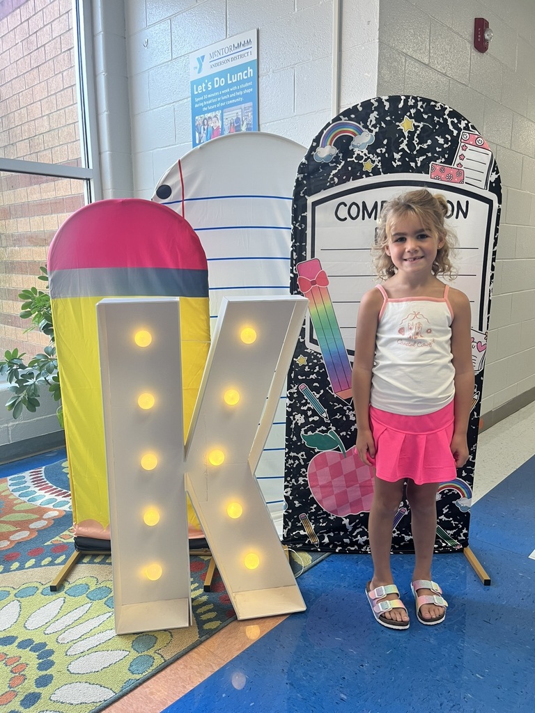 A cheerful kindergarten student smiles brightly while holding a "K" sign surrounded by colorful back-to-school decorations like apples, pencils, and a chalkboard background.