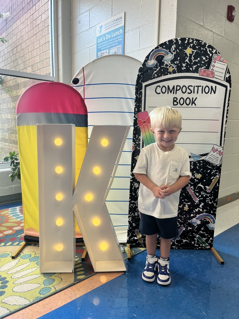 A cheerful kindergarten student smiles brightly while holding a "K" sign surrounded by colorful back-to-school decorations like apples, pencils, and a chalkboard background.