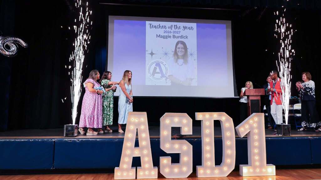 Anderson School District One celebrates 2026–2027 Teacher of the Year Maggie Burdick on stage as colleagues applaud. Spark fountains frame the stage, with illuminated “ASD1” letters in front and her Teacher of the Year recognition displayed on the screen behind her.