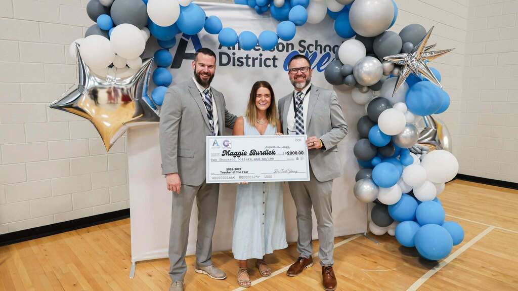 Three school district staff members pose together in front of a blue, white, silver, and gray balloon display. The woman in the center, Maggie Burdick, holds an oversized $2,000 check recognizing her as the 2026–2027 Teacher of the Year.