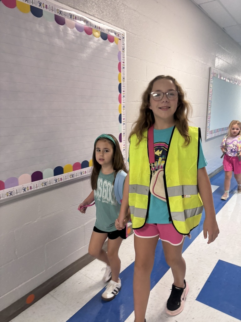 Three elementary students walk through a school hallway. One student wears a bright yellow safety vest while holding hands with a younger student wearing a backpack.